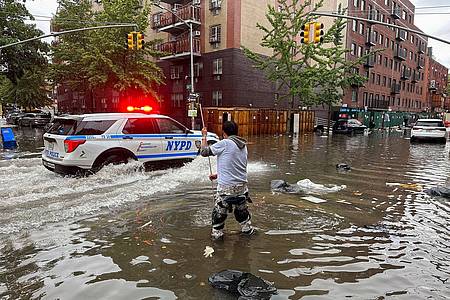 Hochwasser auf den Straßen von New York: Im Stadtteil Brooklyn versucht ein Mann, mit einem  Stock einen Abfluss zu reinigen, während ein Polizeiwagen vorbeifährt.